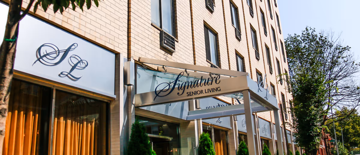 Exterior view of a multi-story brick building with a glass entrance canopy displaying the sign 'Signature Senior Living'. The building has several windows and is surrounded by trees and greenery.