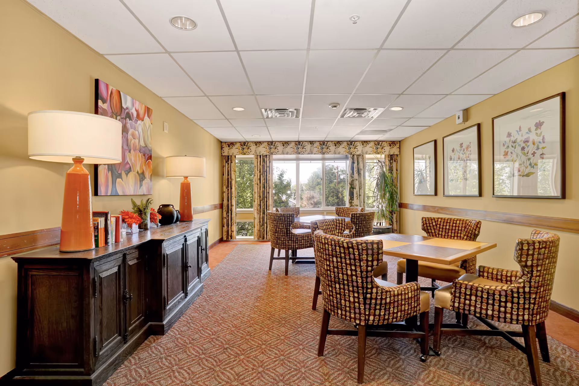 A well-lit interior room with a wooden sideboard on the left holding two orange lamps, books, and decorative items. The room has patterned carpet flooring, a table with four patterned chairs in the foreground, and additional seating near large windows with floral curtains letting in natural light. The walls are adorned with framed floral artwork.