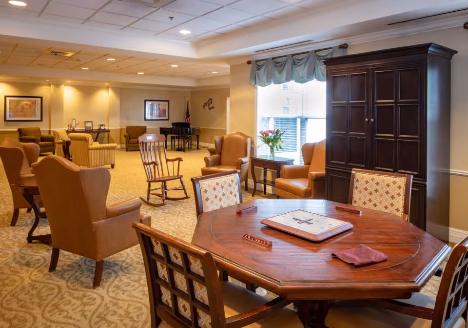 A spacious senior living common area with a large wooden octagonal table in the foreground set up for a board game. Surrounding the table are cushioned chairs with patterned upholstery. In the background, there are several comfortable armchairs and a rocking chair arranged in small groupings. A piano and framed artwork are visible on the far wall, and a window with a valance lets in natural light. The room has warm lighting and carpeted floors.