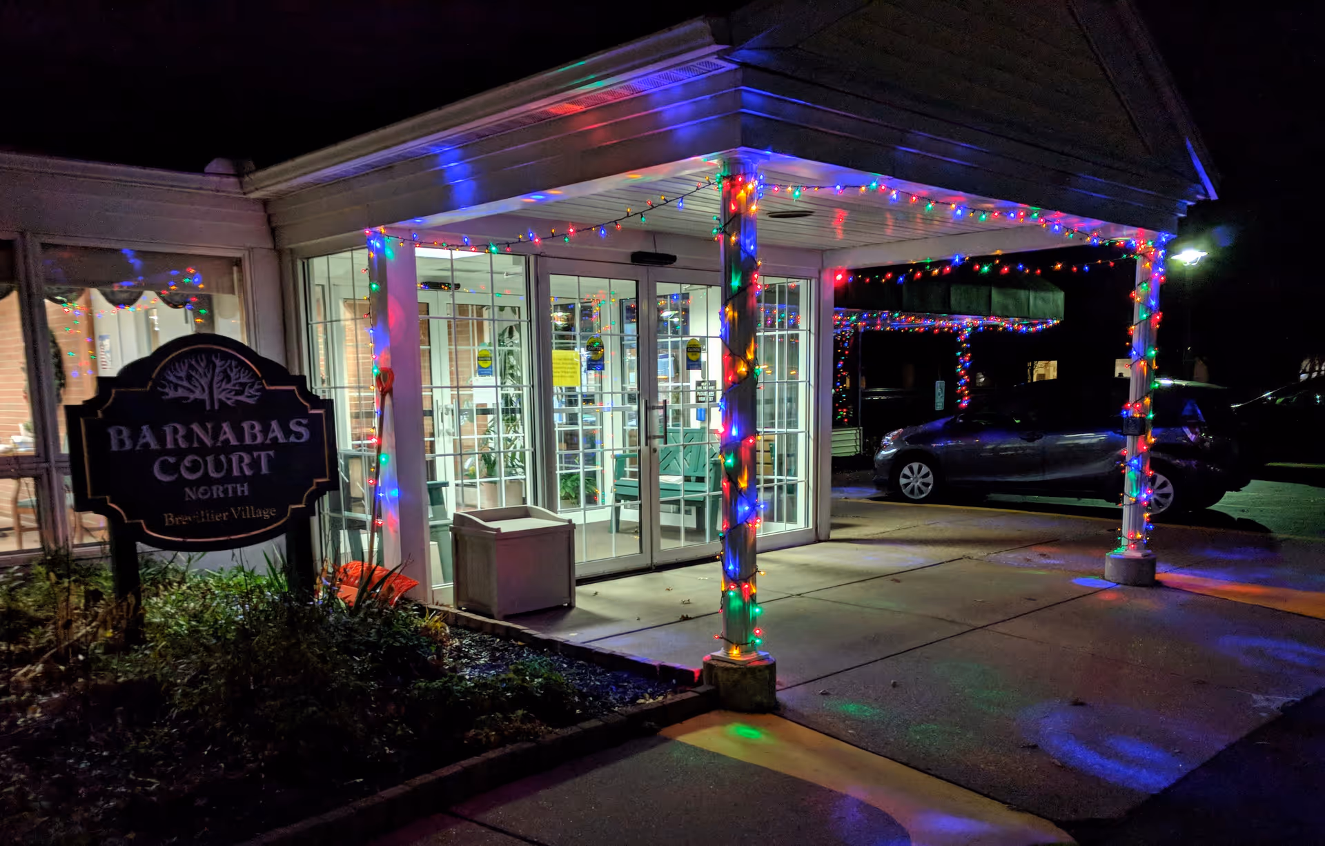 Entrance to Barnabas Court North at Brevillier Village decorated with colorful string lights at night. The entrance has glass doors and a covered walkway with pillars wrapped in lights. A sign for Barnabas Court North is visible on the left side near some plants. A car is parked nearby in the dark.