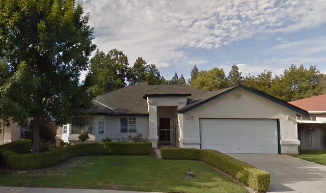 Front exterior view of a single-story house with a two-car garage, a driveway, and well-maintained green lawn and hedges. There are several trees in the background under a partly cloudy sky.