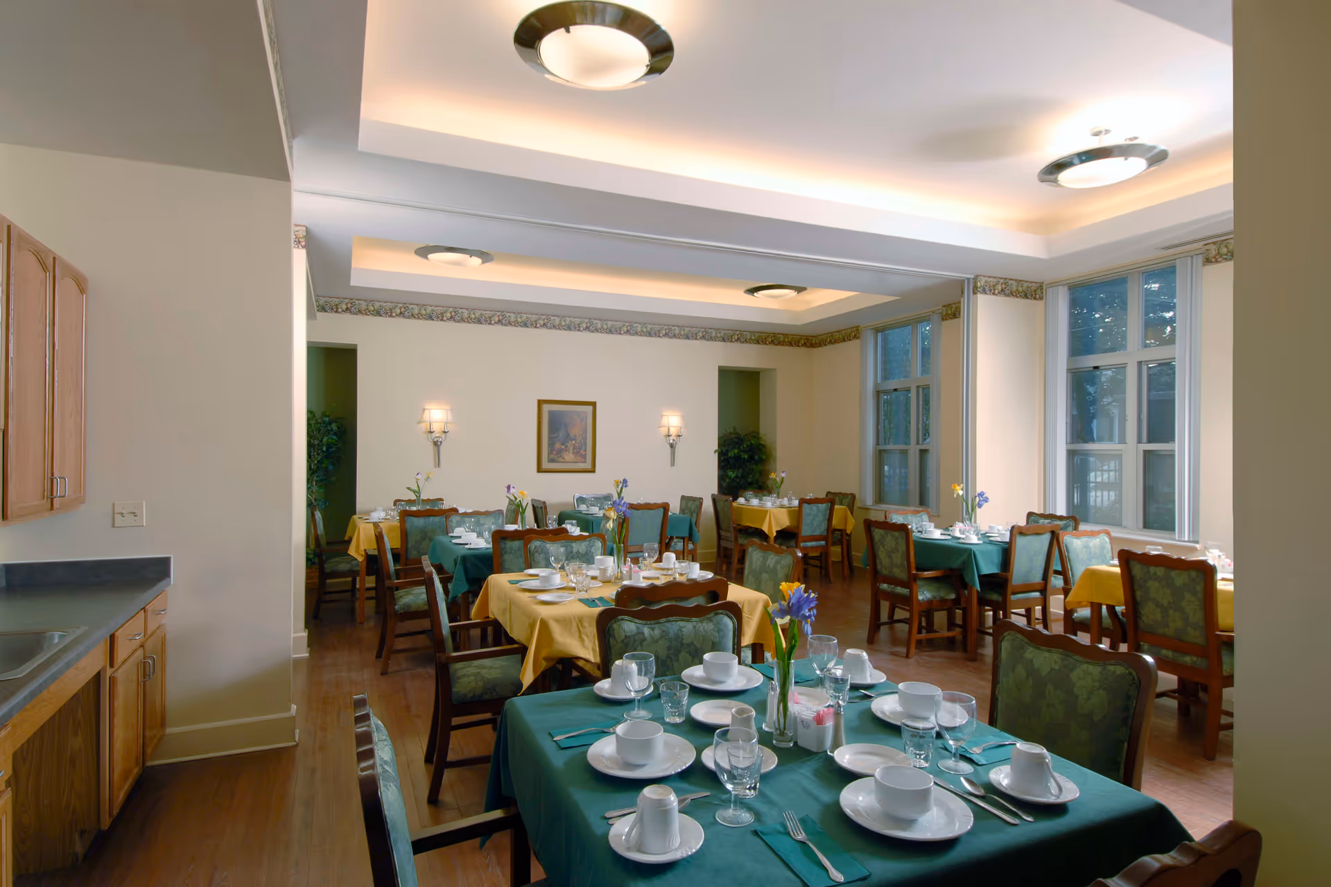 A dining room in Churchview Supportive Living with several tables covered in green and yellow tablecloths, each set with white dishes, cups, glasses, and silverware. The room has wooden chairs with green upholstery, large windows letting in natural light, and soft ceiling lighting. There are plants and framed artwork on the walls.