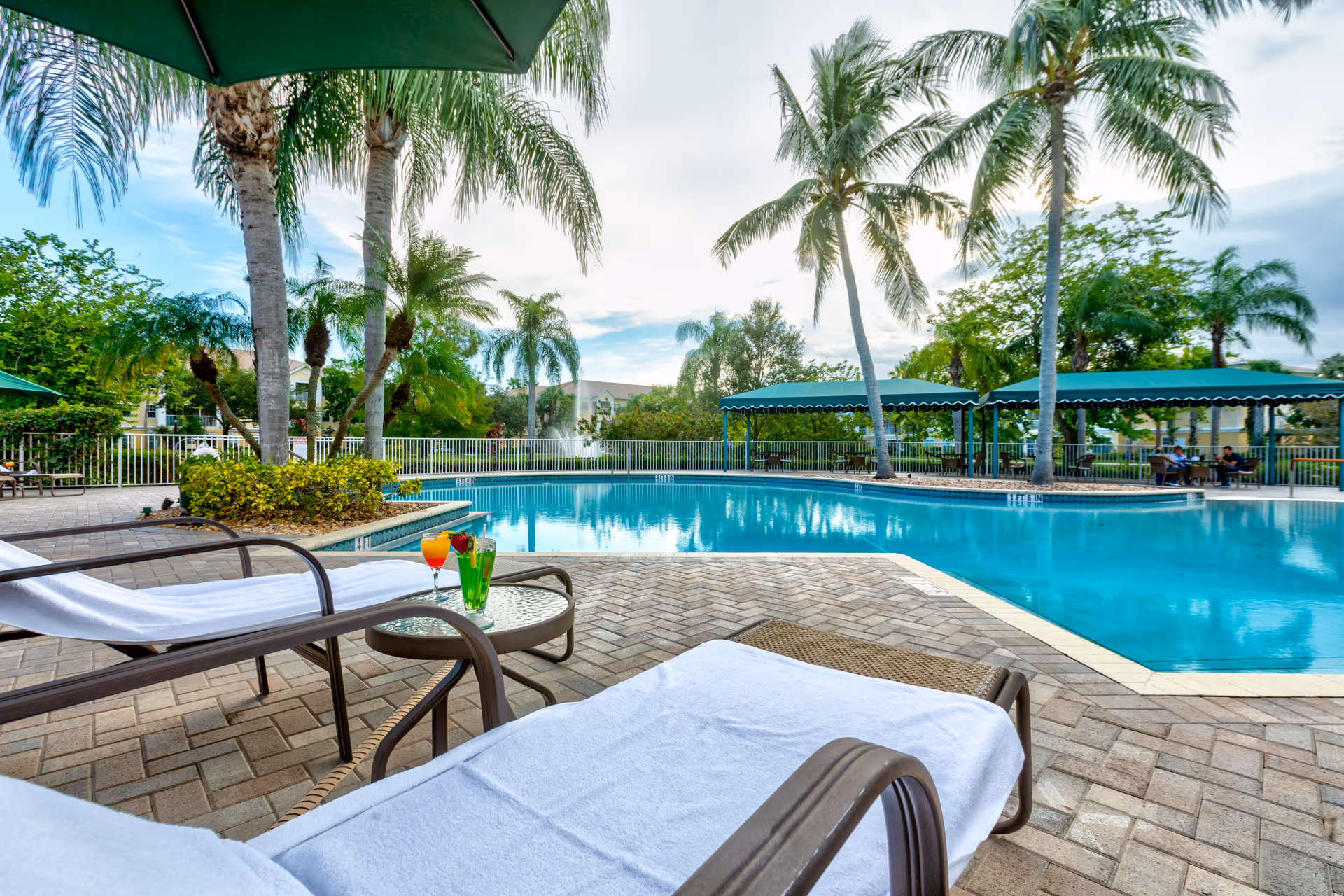 Outdoor swimming pool area with lounge chairs covered with white towels, a small table with colorful drinks, palm trees, and shaded seating areas under green canopies.