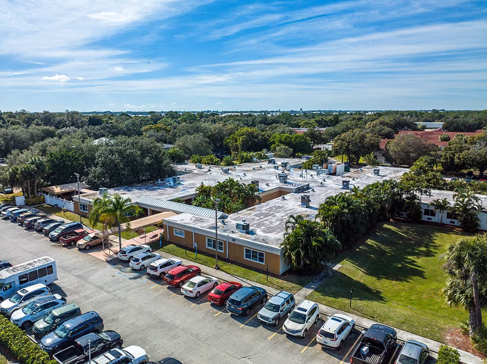 Aerial view of Sea Breeze Rehab And Nursing Center showing a single-story building with a flat roof surrounded by trees and greenery. There is a parking lot with multiple cars parked in front of the building under a partly cloudy blue sky.