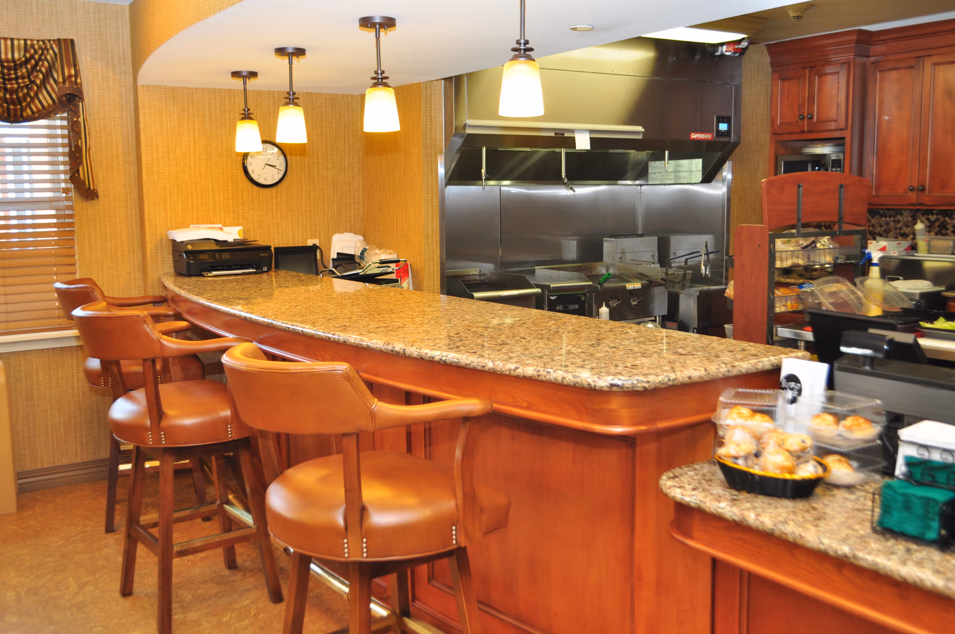 Interior view of a kitchen area with a long granite countertop bar and several brown leather bar stools. The kitchen features stainless steel appliances and cooking equipment in the background, wooden cabinets, and a window with blinds and a valance. There are hanging pendant lights above the bar and a clock on the wall.
