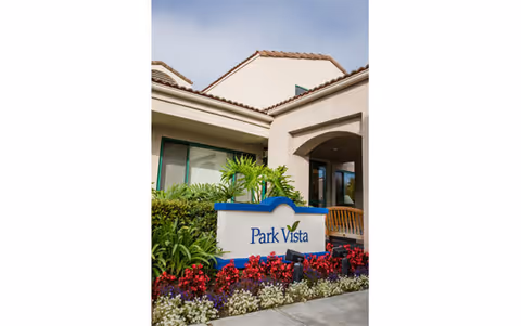 Exterior view of Park Vista Health Center showing the building entrance with a sign that reads 'Park Vista' surrounded by green plants and colorful flowers under a partly cloudy sky.