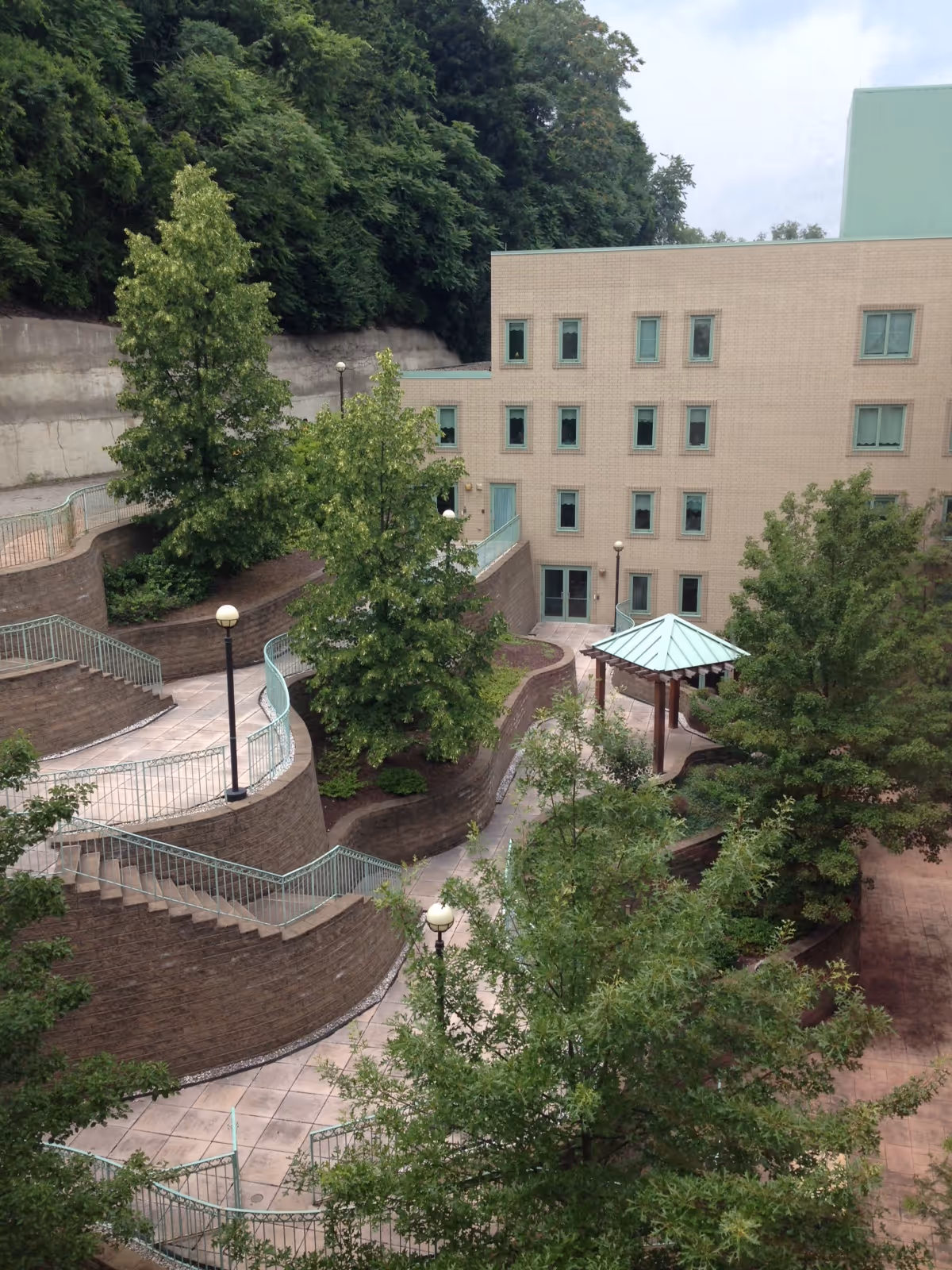 Outdoor courtyard area with winding paved walkways, brick retaining walls, several trees, lamp posts, and a small gazebo with a green roof, adjacent to a multi-story beige building with multiple windows.