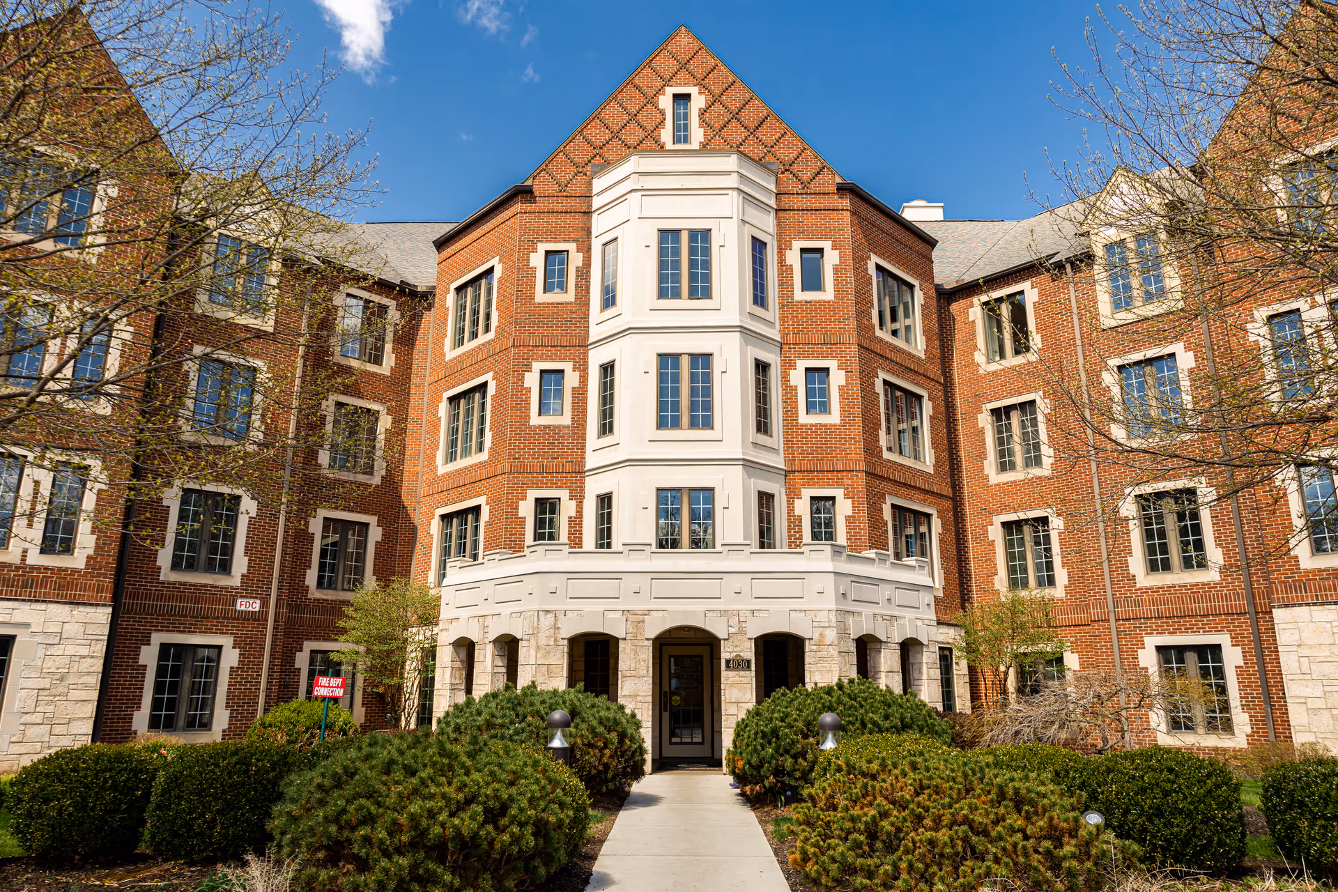 Front exterior view of a multi-story brick building with white stone accents, multiple windows, and a central entrance surrounded by greenery and bushes under a blue sky.