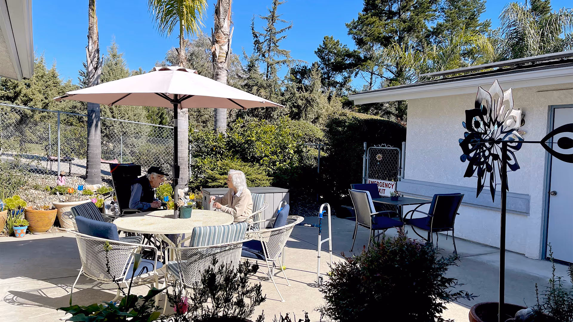 Two elderly individuals sitting at a round outdoor table with an umbrella, surrounded by patio chairs and plants, in a sunny garden area with trees and a white building in the background.