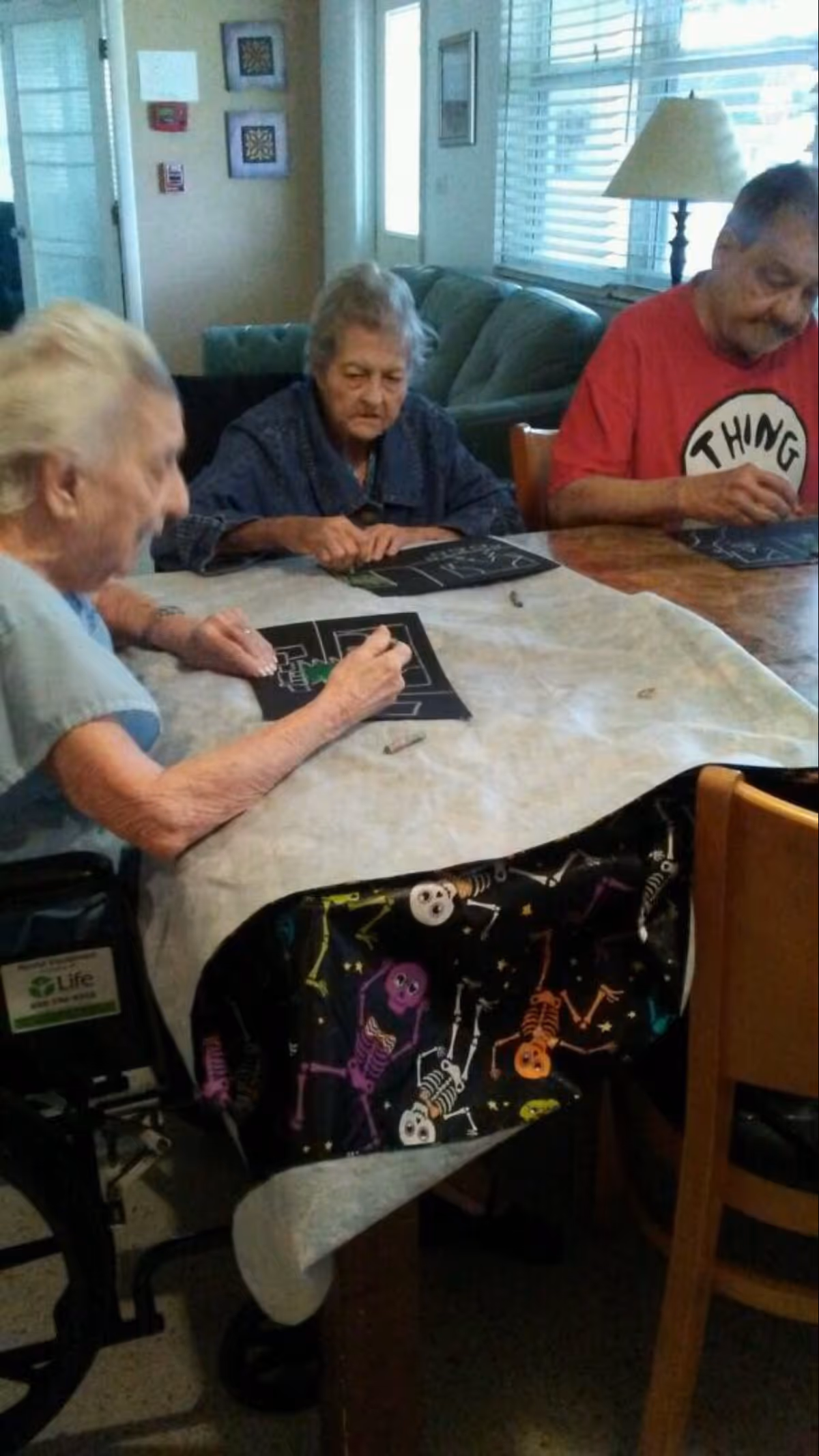 Three elderly individuals sitting around a table engaged in a drawing or coloring activity on black paper. The table is covered with a light-colored tablecloth and a black cloth with colorful skeleton patterns. The room has a window with blinds, a lamp, and a couch in the background.