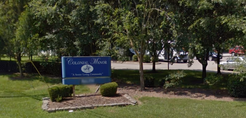 Outdoor view of a blue sign that reads 'Colonial Manor A Senior Living Community' surrounded by grass, small bushes, and trees with a parking lot and several cars in the background.