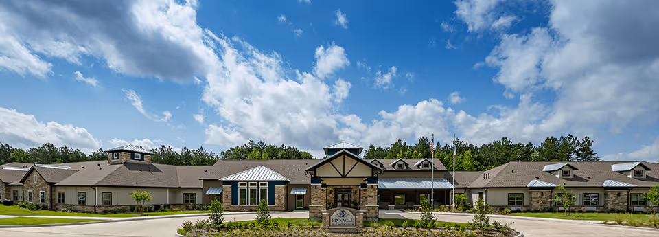 Front exterior view of Pinnacle Senior Living facility with a wide driveway, landscaped greenery, and a partly cloudy blue sky above.