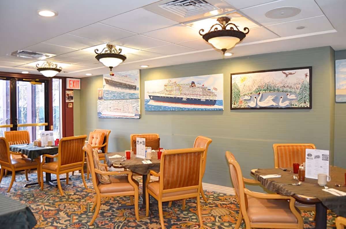 Dining area with several tables covered with dark tablecloths and set with menus, cups, and utensils. The room has wooden chairs with orange upholstery, patterned carpet, and green walls adorned with paintings of ships and swans. Ceiling lights with ornate fixtures illuminate the space, and there is an exit door with a red exit sign.