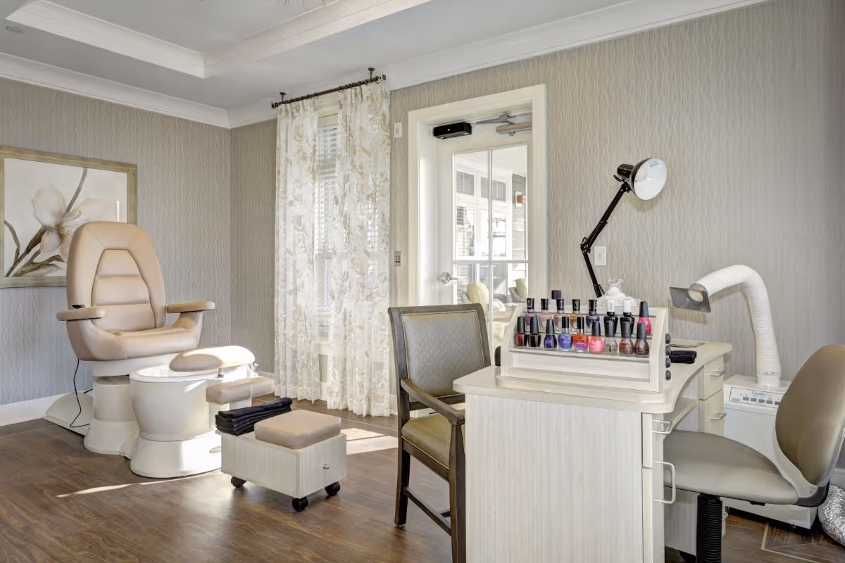 A bright and clean nail salon area with a beige pedicure chair and foot bath on the left, a small rolling stool, and a manicure table on the right with various nail polish bottles displayed. The room has light-colored walls, a window with sheer floral curtains, and a door leading to another area.