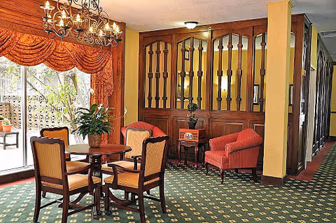 A cozy interior seating area with a wooden table and four upholstered chairs on a green patterned carpet. There are two red armchairs and a small side table with a plant against a wooden partition wall. Large windows with red drapes allow natural light to enter the room.