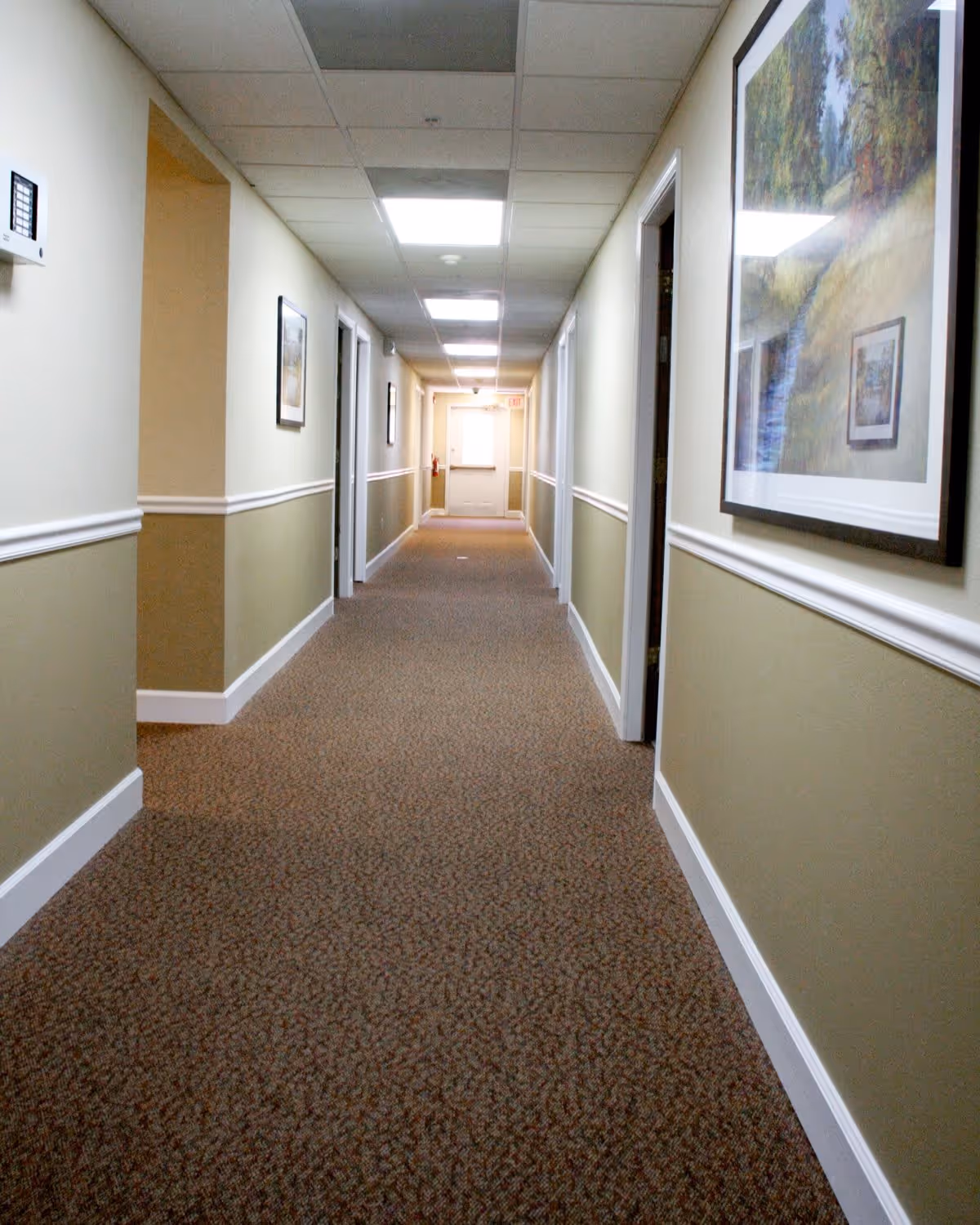 A long, carpeted hallway in a senior living facility with beige and light green walls, white trim, and several framed pictures hanging on the walls. The ceiling has recessed lighting panels, and there are multiple doorways along the hallway.