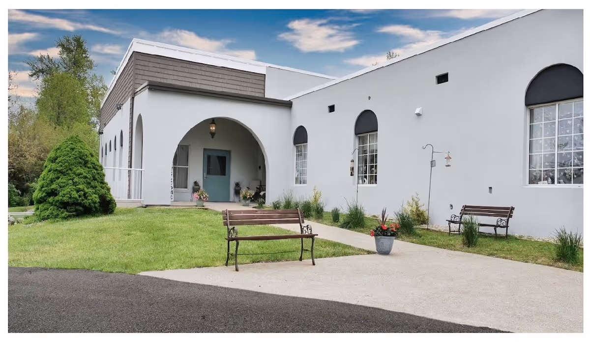 Exterior view of Sunrise Manor Nursing and Rehabilitation building with a white facade, arched entrance, two benches on a concrete path, potted plants, and greenery surrounding the area under a partly cloudy sky.
