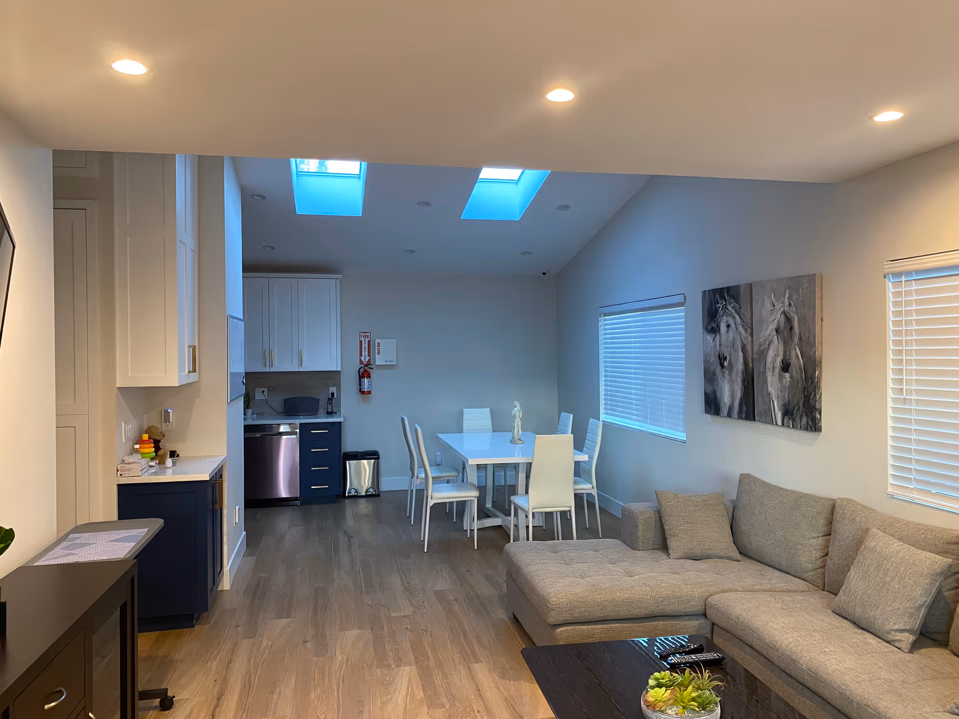 A modern open-concept living and dining area with light wood flooring. The living room has a gray sectional sofa with cushions and a dark coffee table holding a small plant and remote controls. The dining area features a white table with six white chairs under two skylights. The kitchen area has white upper cabinets and dark lower cabinets with a stainless steel dishwasher. Two paintings of horses hang on the wall above the sofa, and windows with blinds are on the right side.