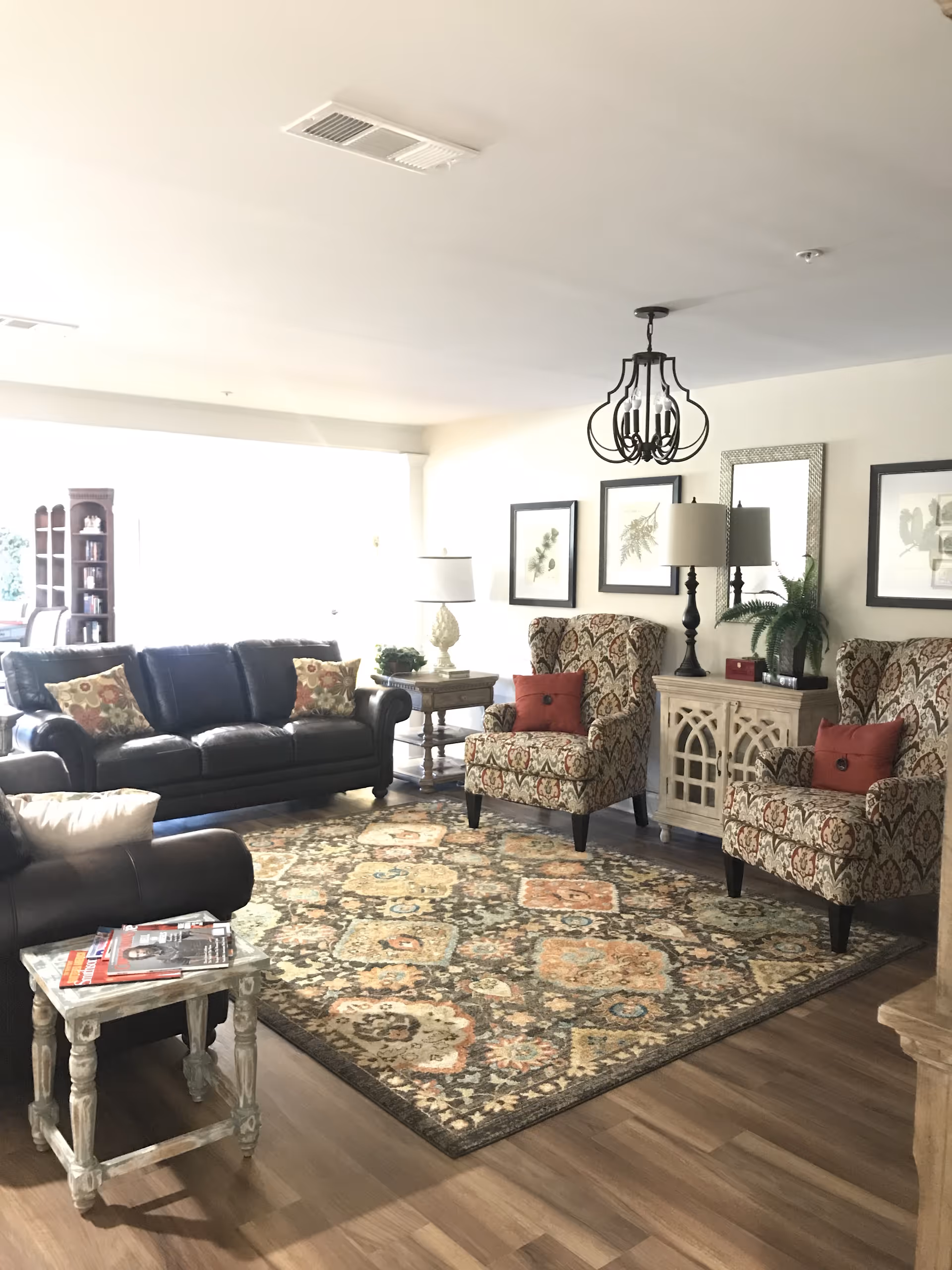 A bright living room with a large patterned area rug on wooden flooring. The room features a dark leather sofa with floral cushions, two patterned armchairs with red cushions, a wooden side table with magazines, and a decorative cabinet with a lamp and plant. Framed botanical prints and a mirror hang on the wall, and a black chandelier is suspended from the ceiling.