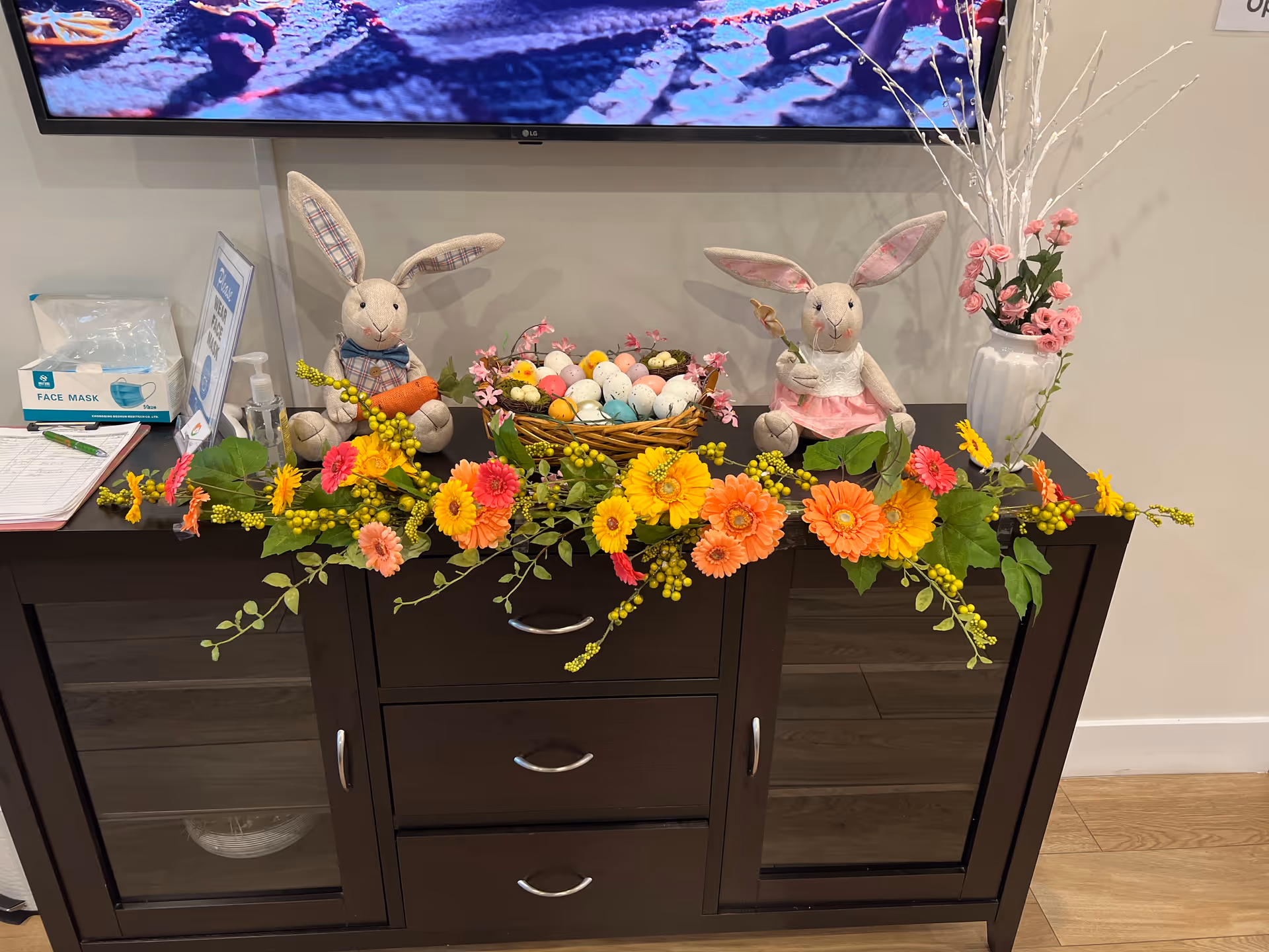 A dark wooden cabinet decorated with colorful artificial flowers, two plush bunny dolls, and a basket filled with pastel-colored Easter eggs. On the left side of the cabinet, there is a box of face masks, a hand sanitizer bottle, and a clipboard with a pen. On the right side, there is a white vase with pink flowers and white decorative branches. A large TV screen is mounted on the wall above the cabinet.
