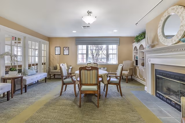 A cozy common area with a round table surrounded by six chairs in the center. The room features a fireplace with a decorative mirror above it on the right, built-in shelves with plants and decor, and a large window with a blue valance letting in natural light. There are additional seating options including armchairs and a bench along the left wall, with beige walls and carpeted flooring.