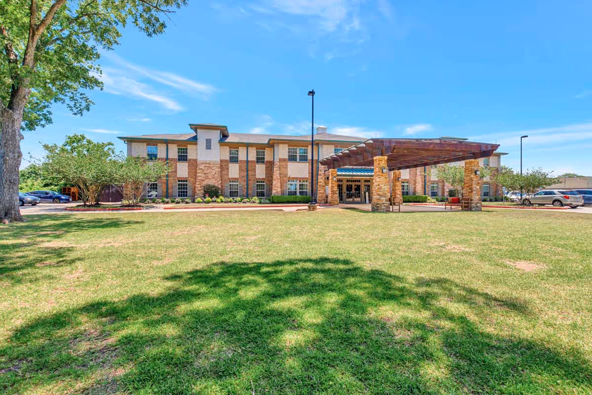 Front exterior view of The Avenues of Fort Bend senior living facility with a large grassy lawn, a covered entrance supported by stone pillars, and a two-story building under a blue sky.