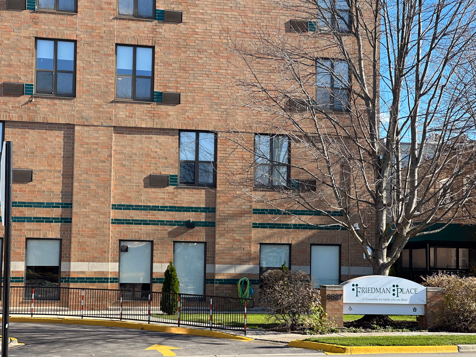 Exterior view of a brick building with multiple windows and a leafless tree in front. A sign near the tree reads 'Friedman Place, A Community for Adults who are Blind'. There is a driveway with a yellow arrow and a black metal fence along the sidewalk.