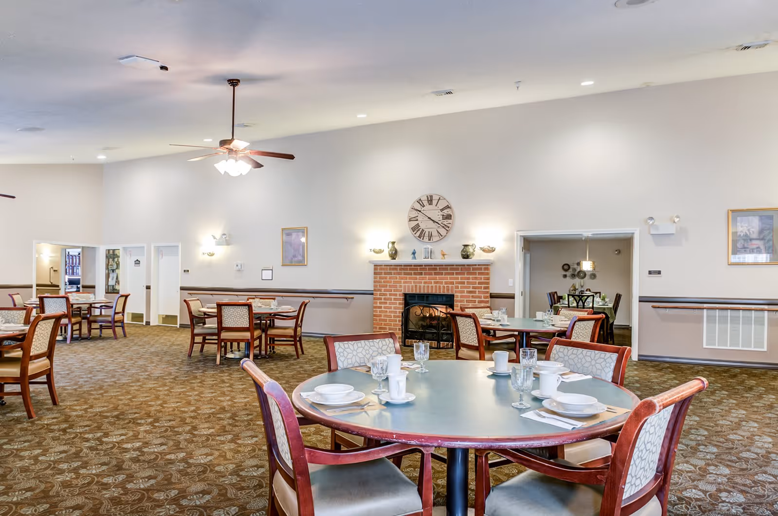 A spacious dining room with multiple round tables set with white dishes, cups, and glasses. The room features a brick fireplace with a large clock above it, wall sconces, framed artwork, and ceiling fans with lights. The carpet has a patterned design, and there are several chairs around each table.