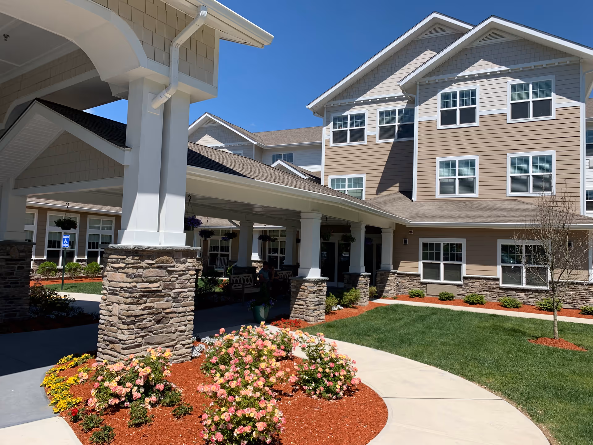 Exterior view of Kennedy Meadows Gracious Retirement Living facility showing a covered entrance with stone pillars, landscaped flower beds with pink and yellow flowers, green lawn, and a multi-story building with beige siding and white trim under a clear blue sky.