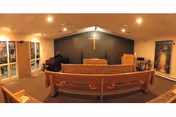 Interior view of a chapel or worship room with wooden pews facing a raised platform with a pulpit and a cross mounted on a dark blue wall. There are stained glass windows on the left side and religious banners on the right side. The room is softly lit with ceiling lights.