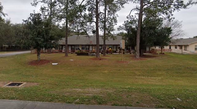 Single-story building with a brown roof surrounded by trees and a grassy lawn, viewed from across a paved road under an overcast sky.