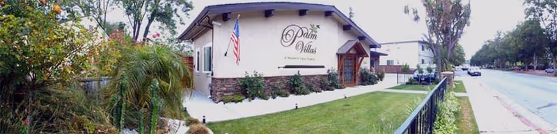 Exterior view of Palm Villas Memory Care Campbell building with an American flag mounted near the entrance, surrounded by greenery and a sidewalk along the street.
