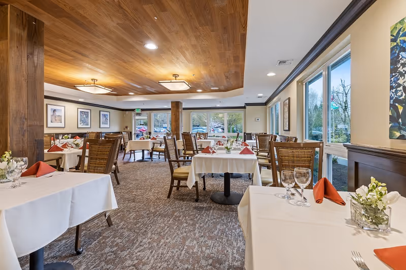 A bright dining room with multiple tables covered in white tablecloths, each set with wine glasses, silverware, and red folded napkins. The room features large windows letting in natural light, wooden ceiling panels, carpeted floor, and framed artwork on the walls.