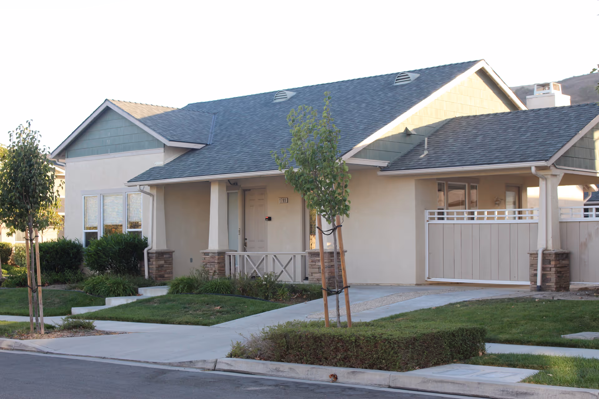 Exterior view of a single-story residential building with a covered porch, small trees, shrubs, and a well-maintained lawn in front. The building has a gray shingled roof and beige walls with stone accents on the porch columns.