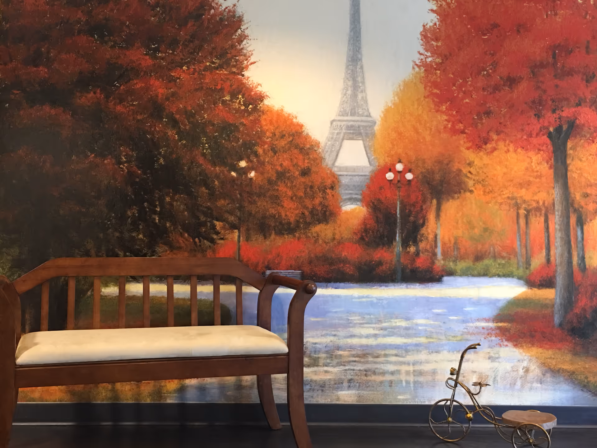 A wooden bench with a cushioned seat is placed indoors in front of a large mural depicting a scenic autumn park with colorful red, orange, and yellow trees, a pathway, and the Eiffel Tower in the background. A small decorative tricycle sculpture is on the floor to the right of the bench.