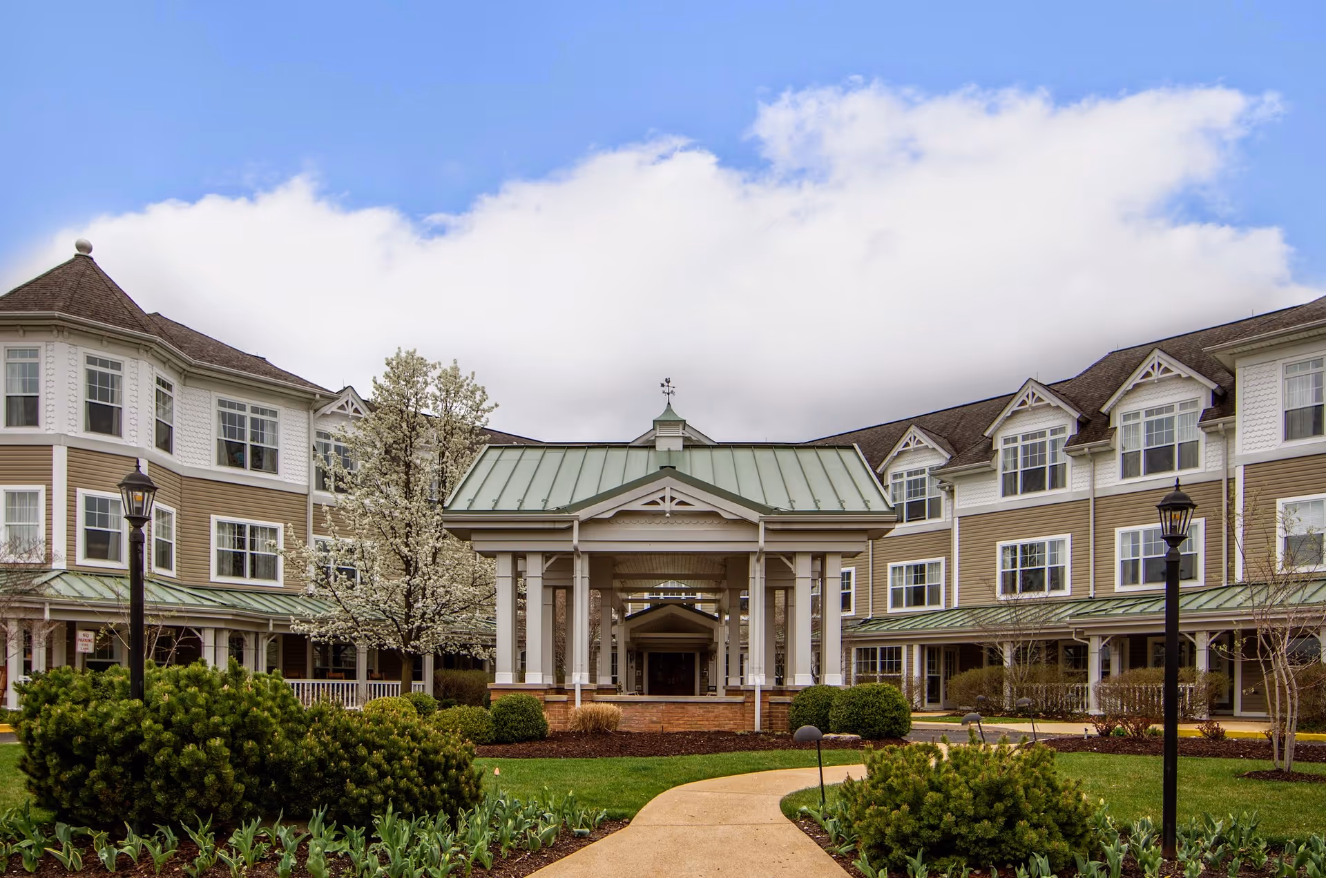 Exterior view of a senior living facility with a covered entrance, manicured bushes, trees, and a pathway leading to the entrance. The building has multiple windows and a green roof with white and beige siding.