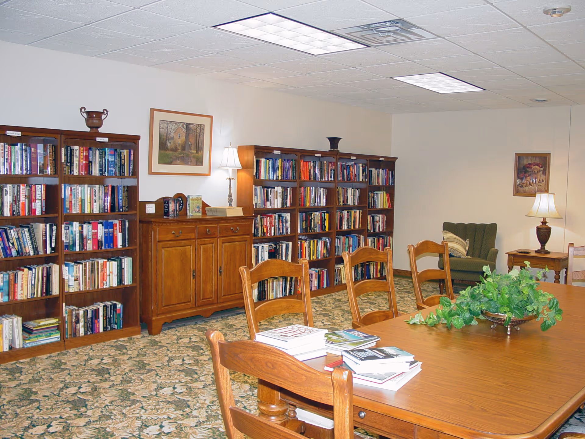 A cozy library room with wooden bookshelves filled with books, a wooden table with chairs around it, and a green armchair in the corner. The room has carpeted flooring with a floral pattern, two table lamps providing warm lighting, and framed artwork on the walls.