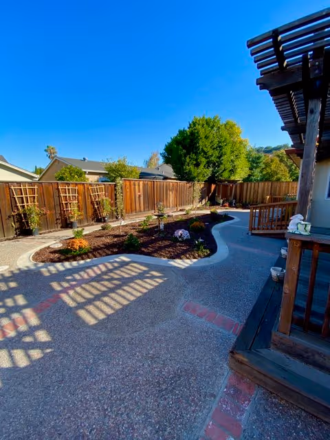 Outdoor garden area with a curved concrete pathway, wooden fence, small plants and shrubs, and a wooden pergola casting shadows on the ground under a clear blue sky.