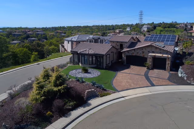 Aerial view of a single-story residential building with a tiled roof and solar panels, situated at the end of a cul-de-sac with landscaped greenery and neighboring houses in the background under a clear blue sky.