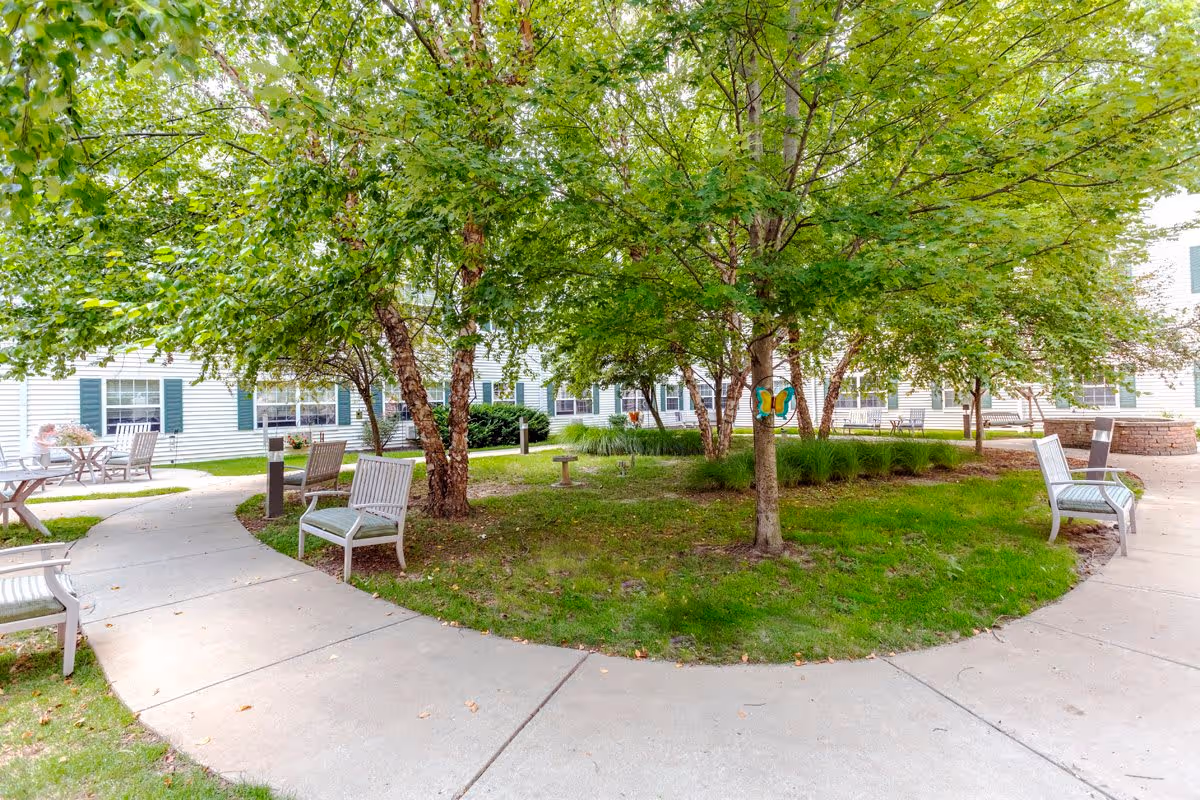 A peaceful outdoor courtyard area with a curved concrete walkway surrounded by green grass and trees. Several benches and small tables with chairs are placed along the path. The background shows a white building with green shutters and multiple windows.