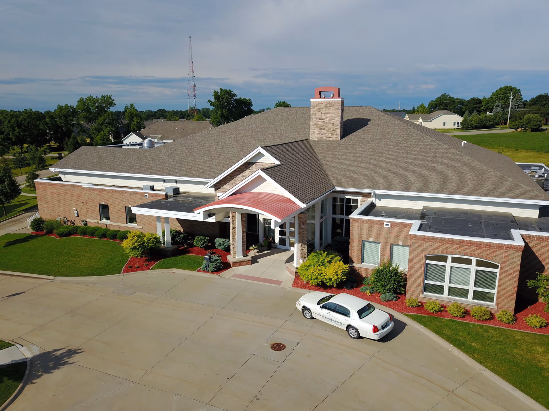 Aerial view of a single-story brick building with a large gray shingled roof and a covered entrance with a red curved awning. The building is surrounded by well-maintained green lawns, shrubs, and trees. A white car with the word 'Fountains' on its side is parked near the entrance. The sky is partly cloudy.