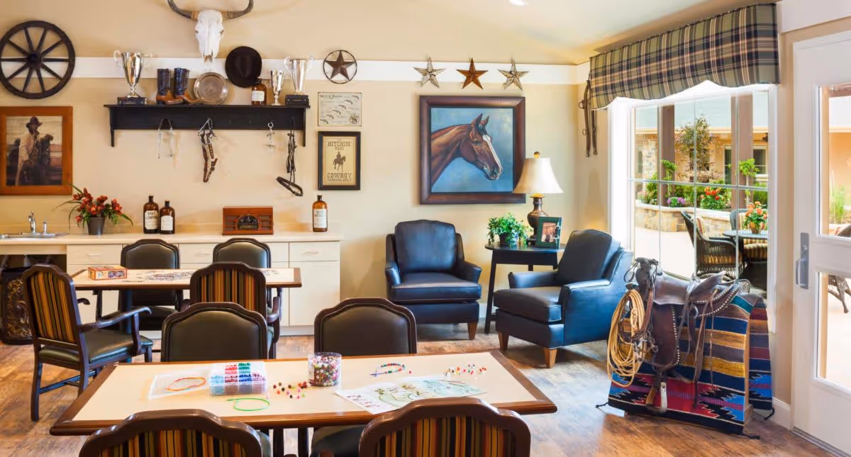 A cozy common area in El Rio Memory Care Community featuring a table with chairs and craft supplies, two black armchairs with a side table and lamp, western-themed decor including a horse painting, cowboy hats, boots, and a saddle, and a large window showing an outdoor patio with plants and seating.