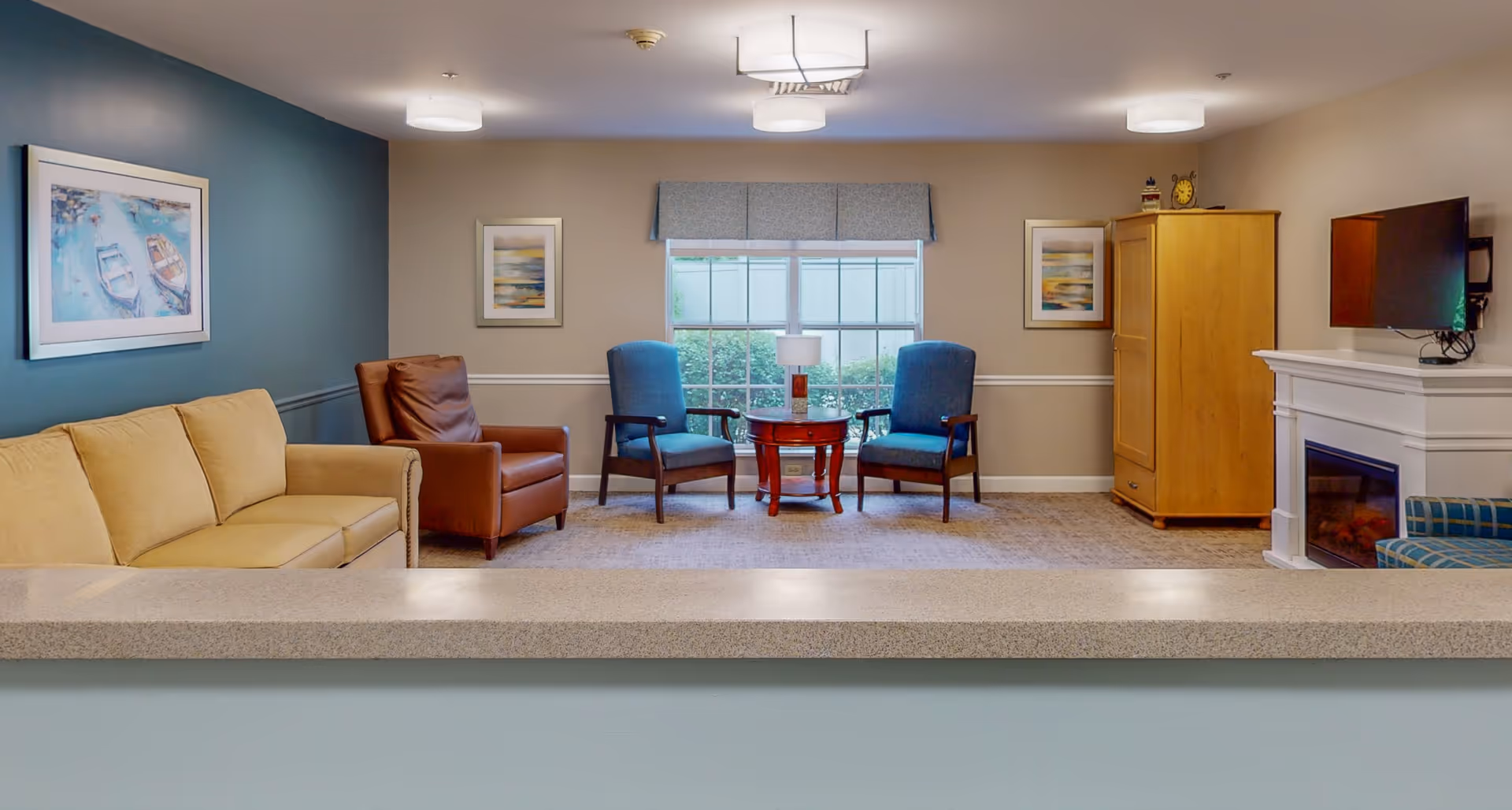 A cozy living room area with a beige sofa, a brown leather armchair, two blue upholstered chairs around a small wooden table with a lamp, a wooden cabinet, a wall-mounted TV above a white fireplace, and framed artwork on the walls. A large window with a valance lets in natural light.