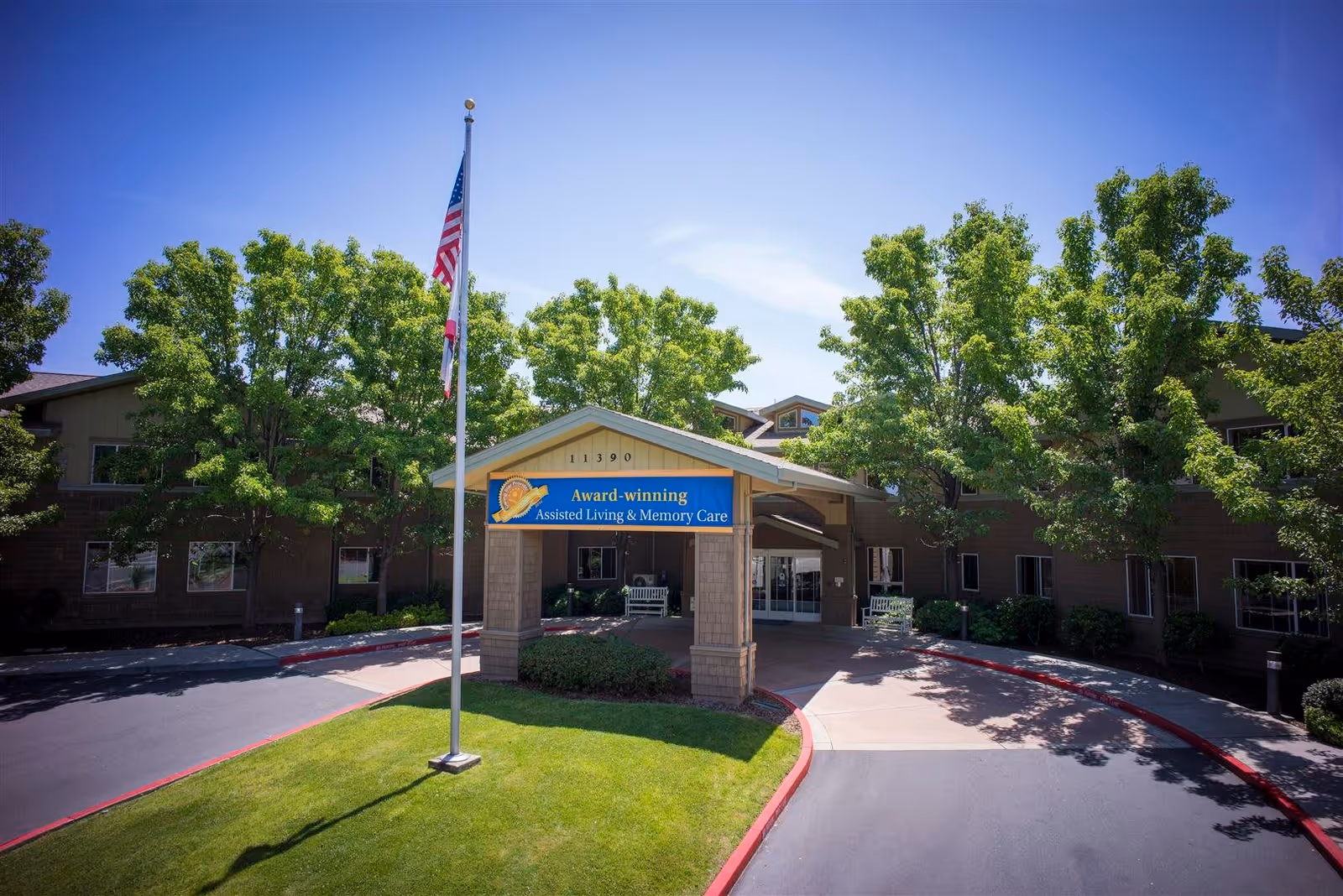 Front exterior view of Eskaton Gold River facility with a covered entrance, an American flag on a flagpole, green trees, and a clear blue sky.