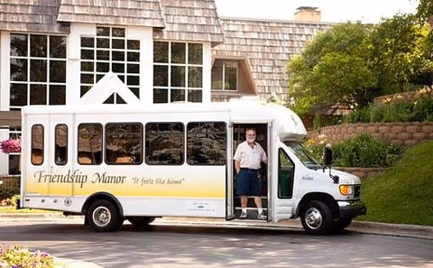 A white shuttle bus with the words 'Friendship Manor' and the slogan 'It feels like home' parked outside a building with large windows and wooden shingles. A man in a white shirt and dark shorts stands at the open door of the bus.