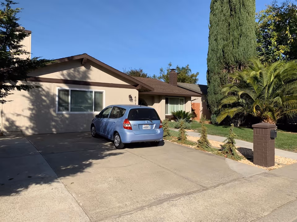 Exterior view of a single-story residential building with a light beige facade and brown roof trim. A blue compact car is parked in the concrete driveway. The front yard features a tall cypress tree, a palm plant, and small shrubs along a stone-lined path leading to the entrance. The sky is clear and blue.