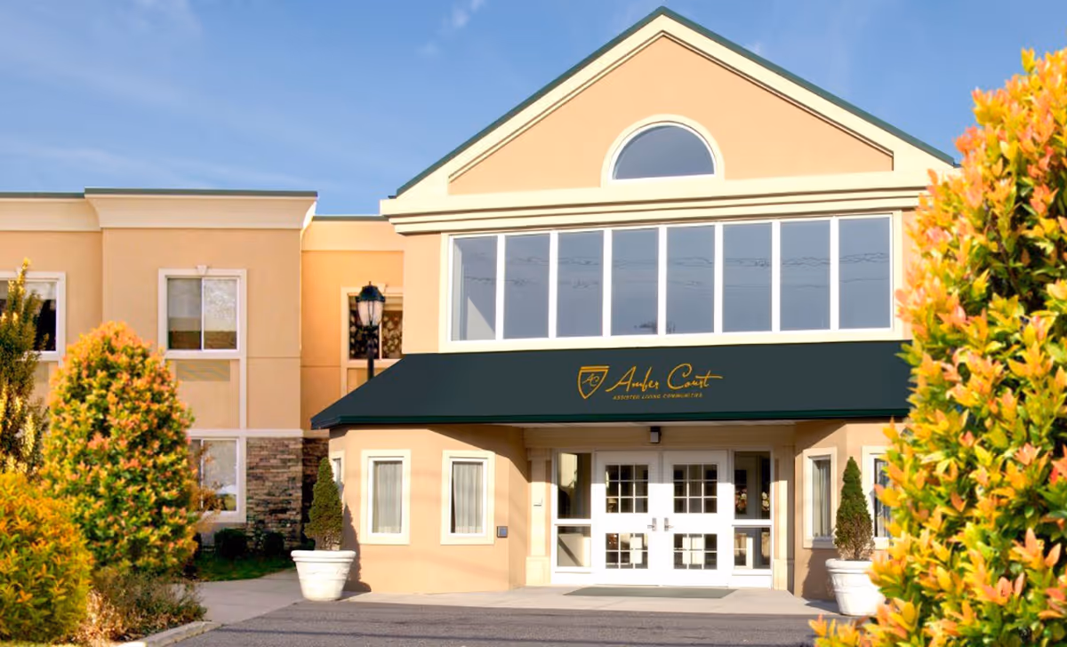 Front exterior view of Amber Court of Westbury assisted living community building with beige walls, large windows, a black awning with the facility's name and logo, and landscaped bushes on either side of the entrance.