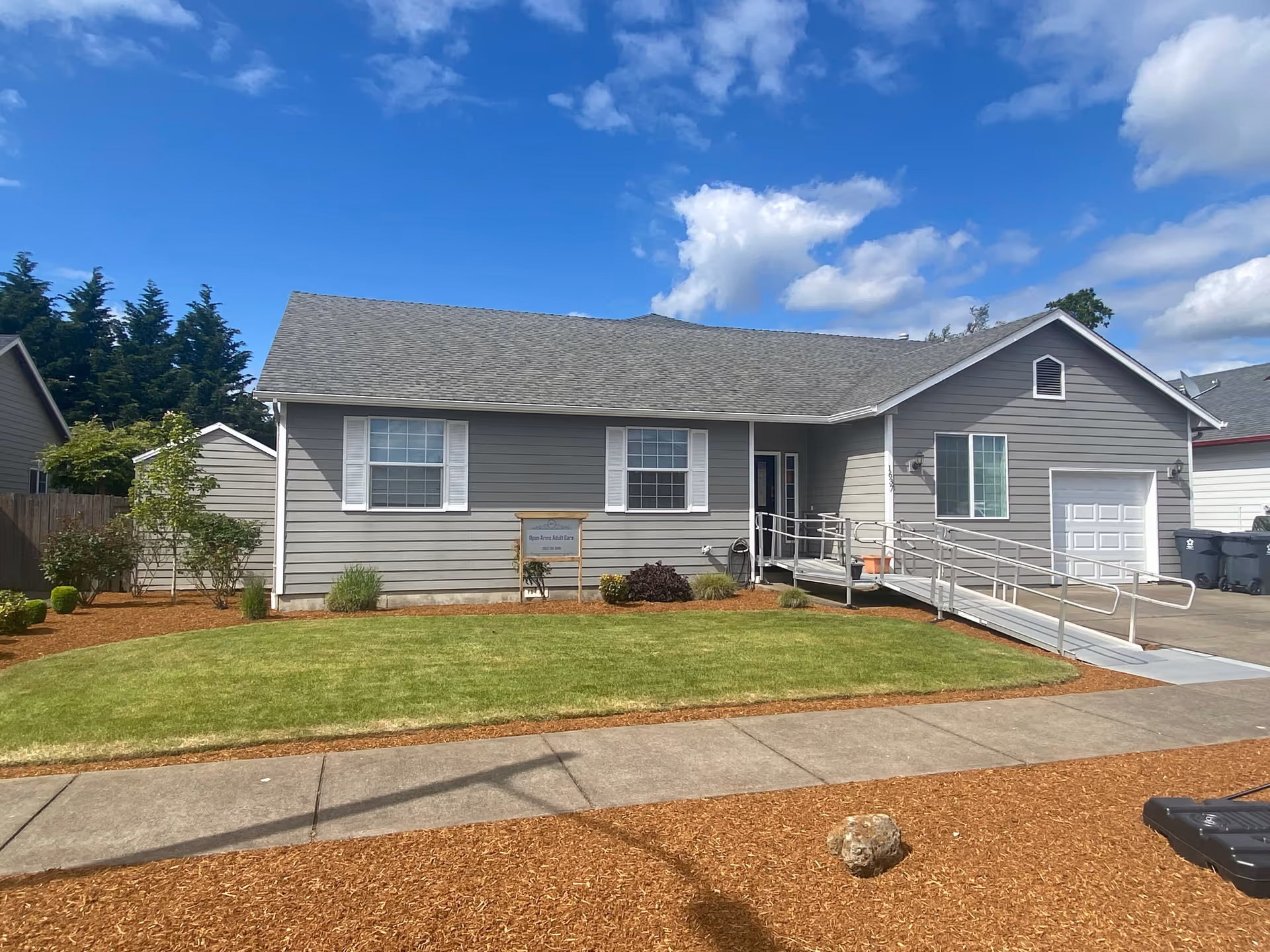 Single-story gray building with white trim and a wheelchair accessible ramp leading to the front door. The building has a well-maintained lawn and some small bushes in front. A sign near the center window reads 'Open Arms Adult Care'. The sky is blue with scattered clouds.