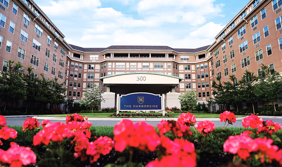 Front exterior view of The Harborside senior living facility with a large building featuring multiple windows, a covered entrance with the number 300 above it, and a sign displaying the facility's name. Bright pink flowers and green shrubs are in the foreground under a partly cloudy sky.