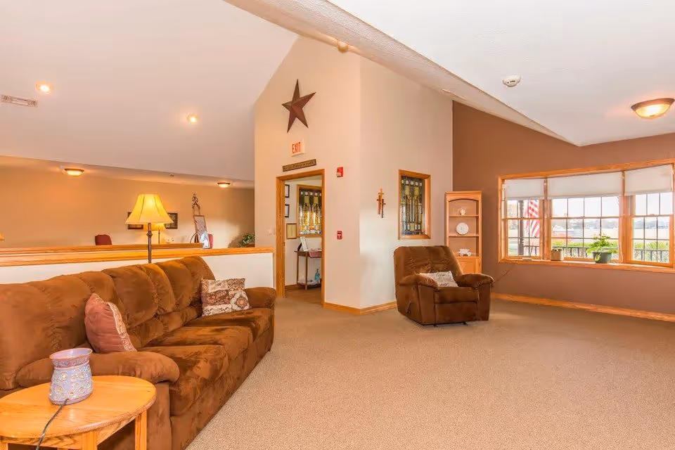 A cozy living room area with a brown sofa and armchair, a wooden side table with a decorative item, a floor lamp, and a large window with a view outside. The walls are painted in neutral tones, and there is a star decoration above a doorway leading to another room.